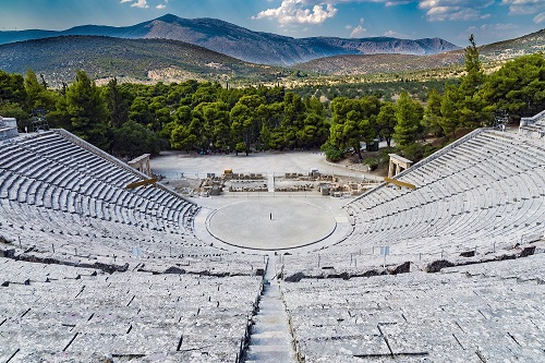 A high shot of an amphitheater made out of stone with green trees and mountains in the background